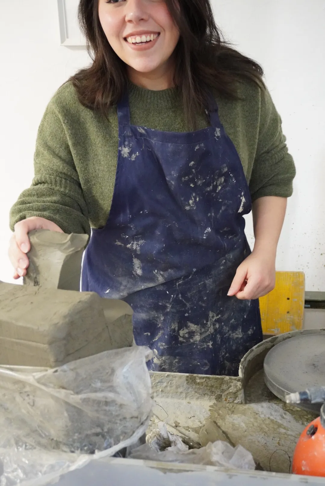 Frida smiles in the studio holding a slab of clay, her apron stained