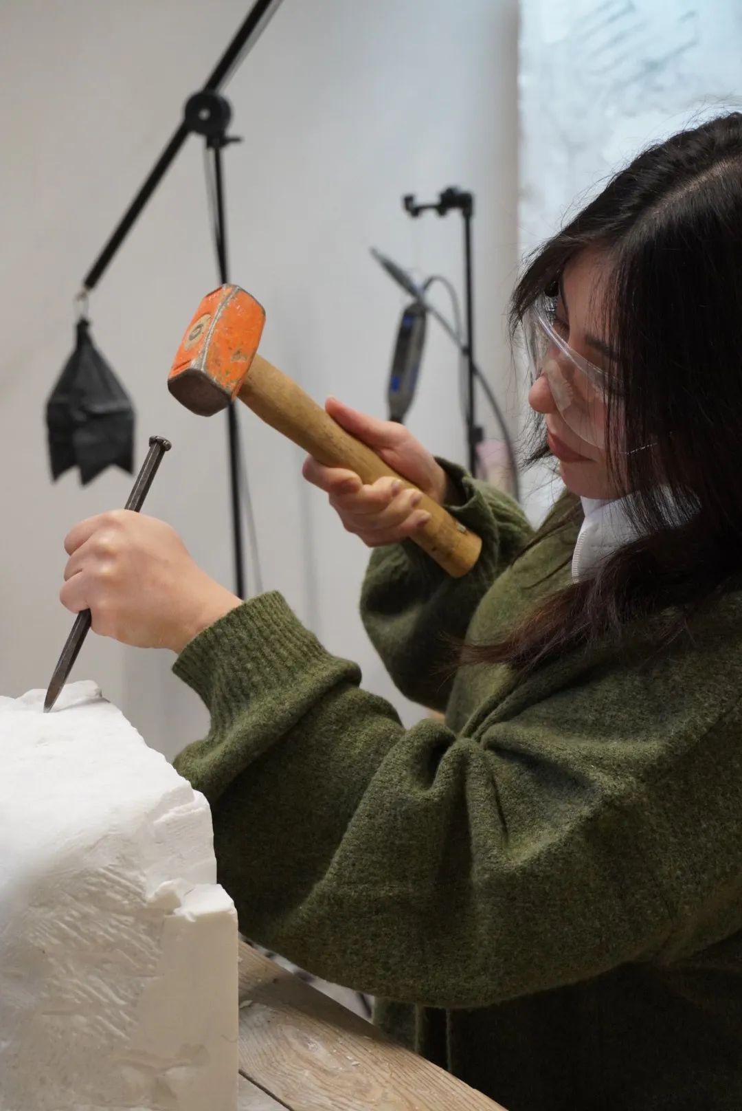 Frida carves a block of marble with a mallet and chisel, wearing safety goggles
